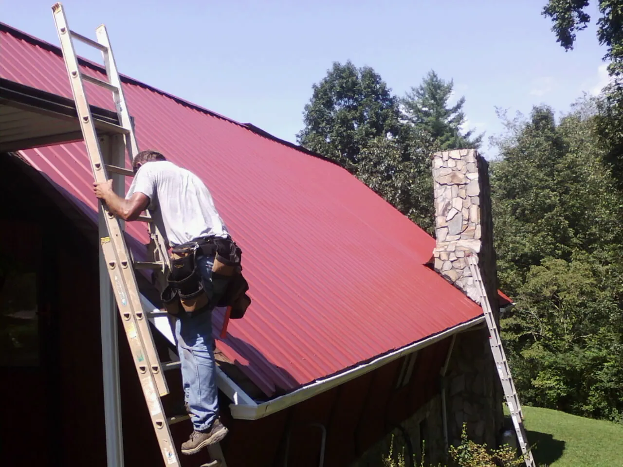 Roofer on a ladder beside a red metal roof and stone chimney — Soffit & Fascia Repair by Roofing Brothers in Canyon Dam
